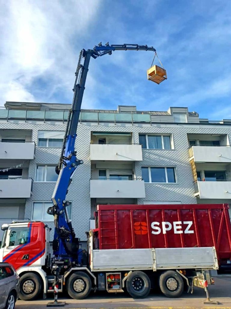 Ein Lastwagenkran der Firma SPEZ hebt eine Holzpalette vor einem mehrstöckigen Wohngebäude in die Luft.